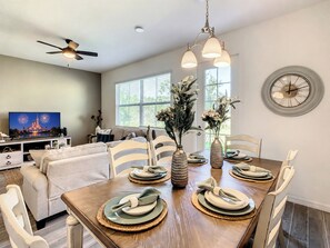 A dining area with a wooden table set for four, surrounded by white chairs. There are two vases with flowers on the table, and a ceiling fan above.