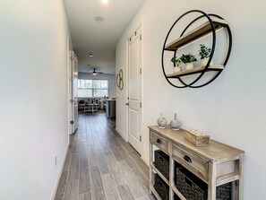 A hallway with a wooden floor, a console table with baskets, a wall-mounted shelf with plants, and a ceiling fan.
