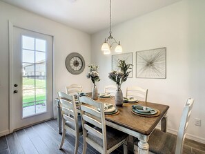 A dining area with a wooden table, white chairs, a vase with flowers, a wall clock, and abstract wall art.