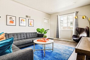 Ein modernes Wohnzimmer mit einem grauen Ecksofa, einem runden Couchtisch und einem großen Fenster mit Vorhängen.