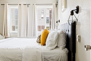 A bedroom with a bed, white bedding, a headboard, a wall-mounted light fixture, and a window with curtains. - Stylish City Condo | King Beds | Sloans Lake 30Day (Denver)