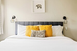 Una cama con cabecera tapizada en gris, ropa de cama blanca y dos almohadas amarillas.