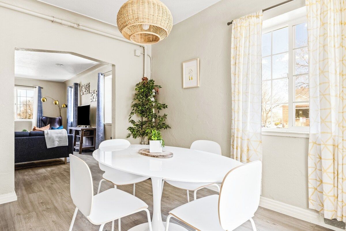 Un coin repas avec une table ronde blanche, des chaises blanches, un luminaire suspendu et une fenêtre avec des rideaux.