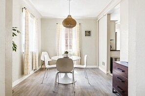 Un coin repas avec une table ronde, des chaises blanches, un luminaire suspendu et une vue sur un mur de briques à travers une fenêtre.