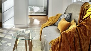 A modern living room with a glass coffee table, a sofa with a mustard blanket, and large windows.