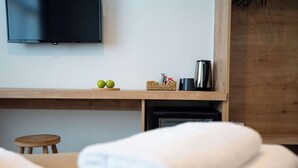 Habitación de hotel con televisor de pantalla plana en la pared, estante de madera con una cesta y cafetera, un taburete pequeño y toallas dobladas con esmero.
