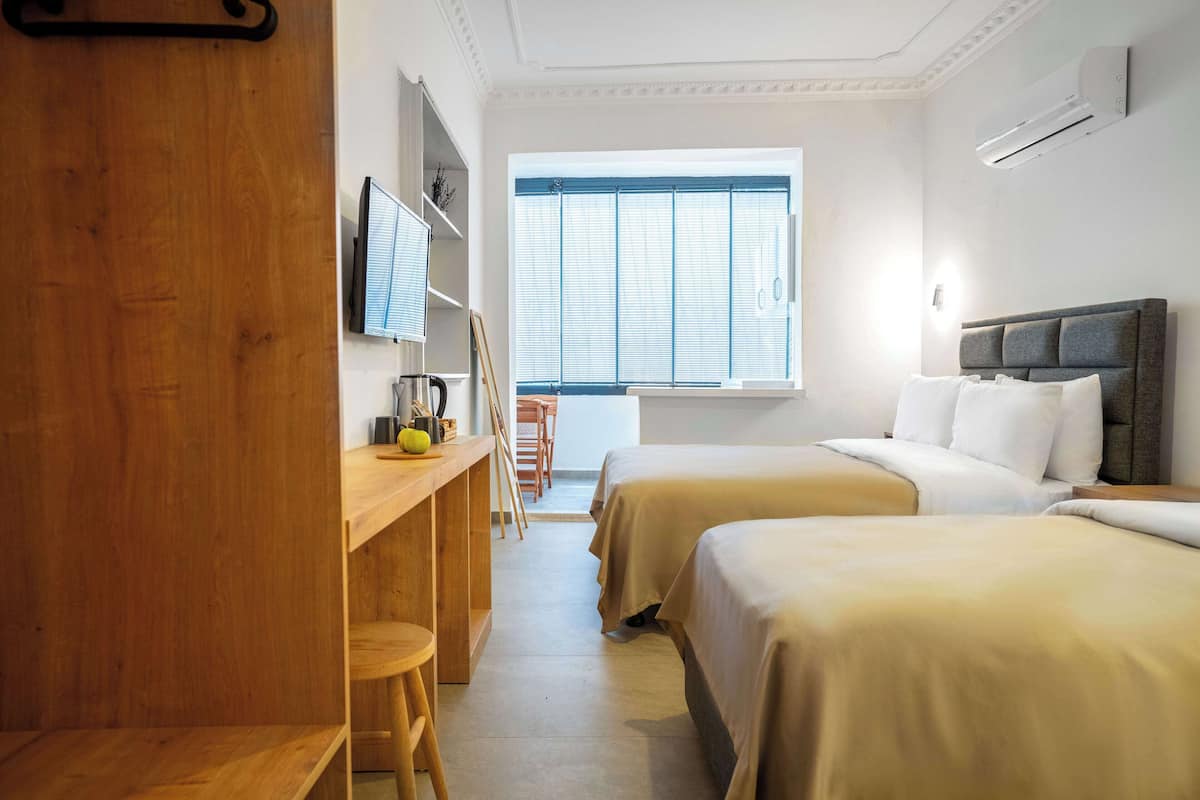 Ein Hotelzimmer mit zwei Betten, einem Schreibtisch, einem Stuhl, einem Fenster mit Jalousien und einer Klimaanlage.