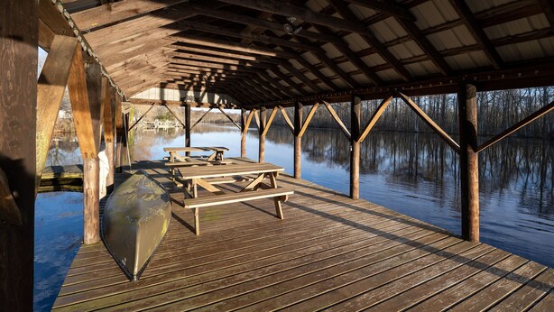 Un muelle de madera con mesas de picnic y un área techada, con vista a una masa de agua tranquila.