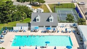 Une piscine entourée de chaises longues et de parasols, avec un bâtiment doté d’un toit en pente et un court de tennis en arrière-plan.