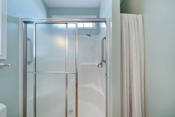 A glass-enclosed shower stall with a white curtain, a visible showerhead, and a white toilet to the left.