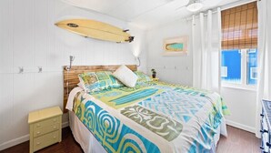 Un dormitorio con cama, una tabla de surf, una ventana con cortinas y una mesita de noche pequeña.