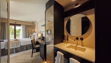 Een moderne hotelkamer met een groot bed, een bureau met stoel, een ronde spiegel en een badkamer met wastafel en handdoekhouder.