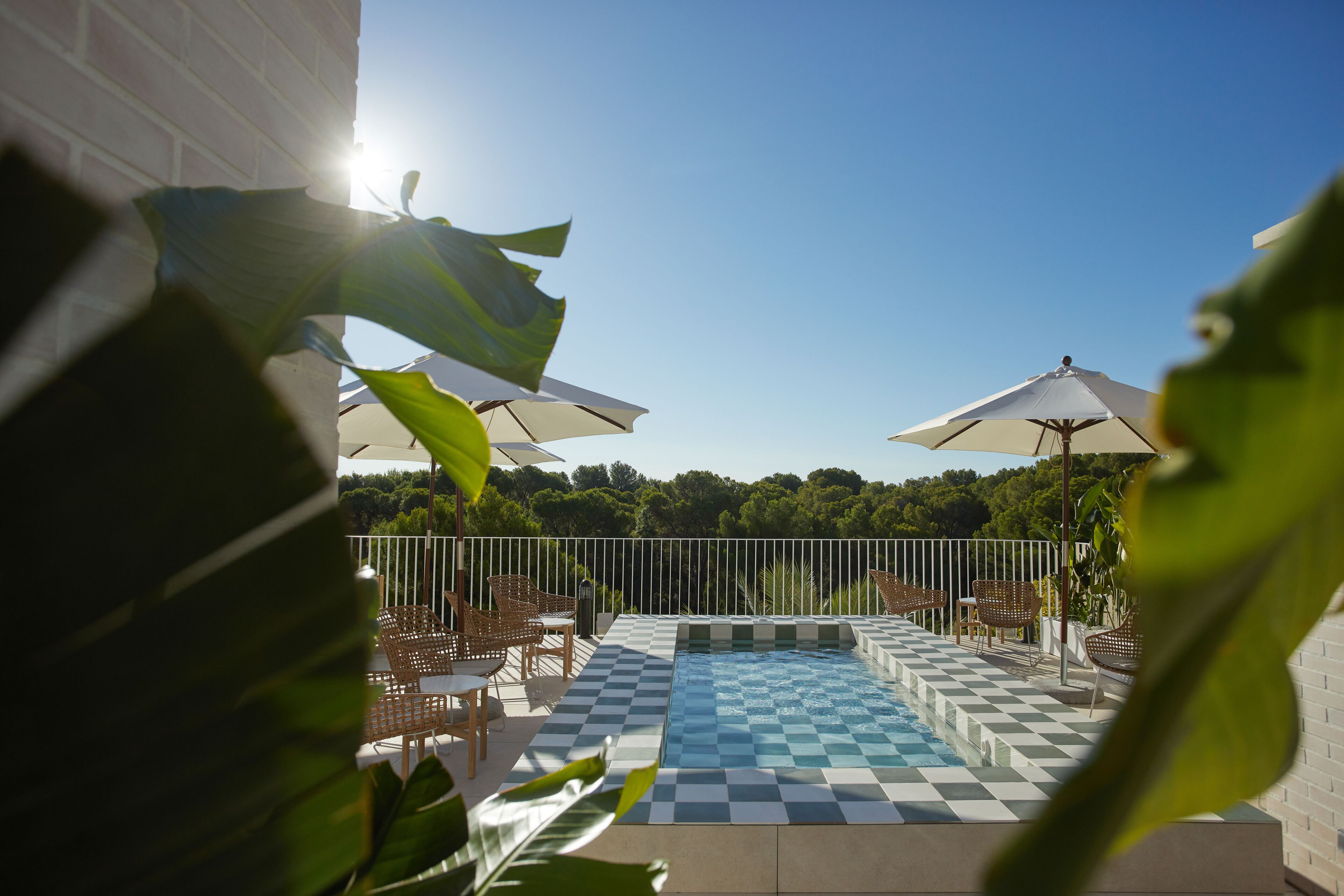 Un espace piscine avec des carreaux aux motifs à damier, entouré de chaises et de parasols, et offrant une vue sur les arbres en arrière-plan.