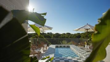 Un espace piscine avec des carreaux aux motifs à damier, entouré de chaises et de parasols, et offrant une vue sur les arbres en arrière-plan.