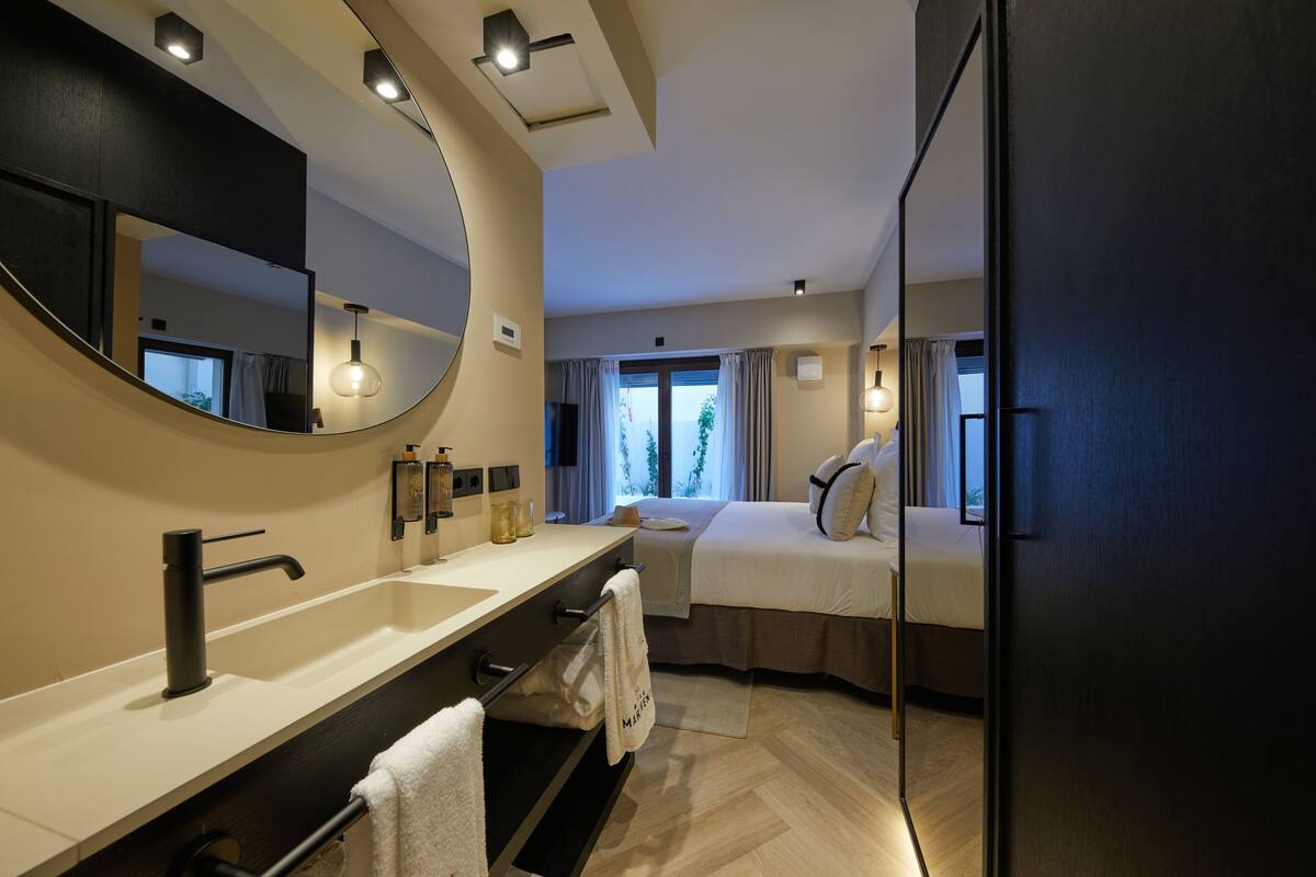 Ein modernes Hotelzimmer mit einem großen Spiegel, einem Doppelbett, einem separaten Badezimmer mit Waschbecken und Ablagefläche sowie Blick ins Freie durch das Fenster.