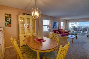 Uma zona de refeições com mesa de madeira e cadeiras amarelas, uma sala de estar com sofá vermelho e uma varanda com mobiliário de exterior.