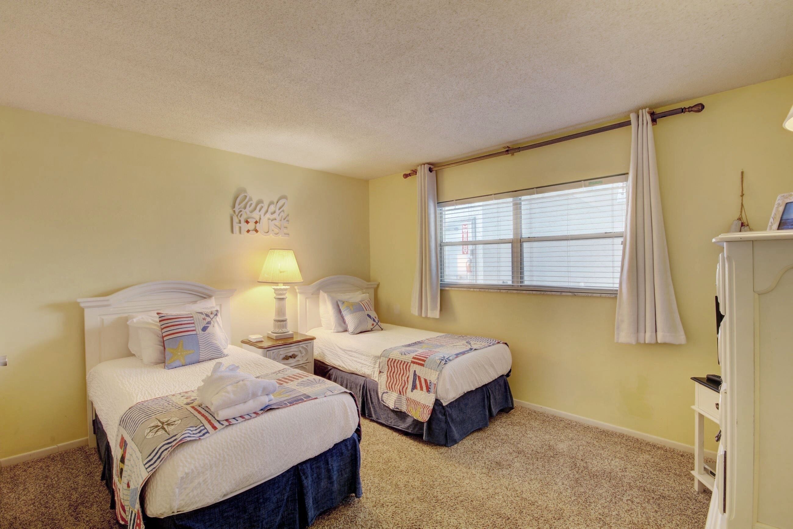 A bedroom with two beds, a nightstand with a lamp, a window with curtains, and a wall decoration.