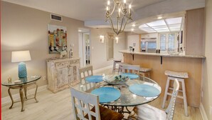 A dining area with a glass table, blue placemats, white chairs, a chandelier, a cabinet, and a painting on the wall.