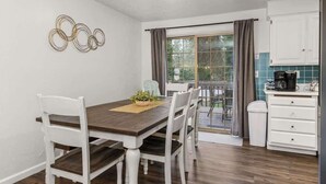 Un comedor con una mesa de madera y sillas blancas, una cocina con muebles blancos y una cafetera, y una ventana con cortinas.