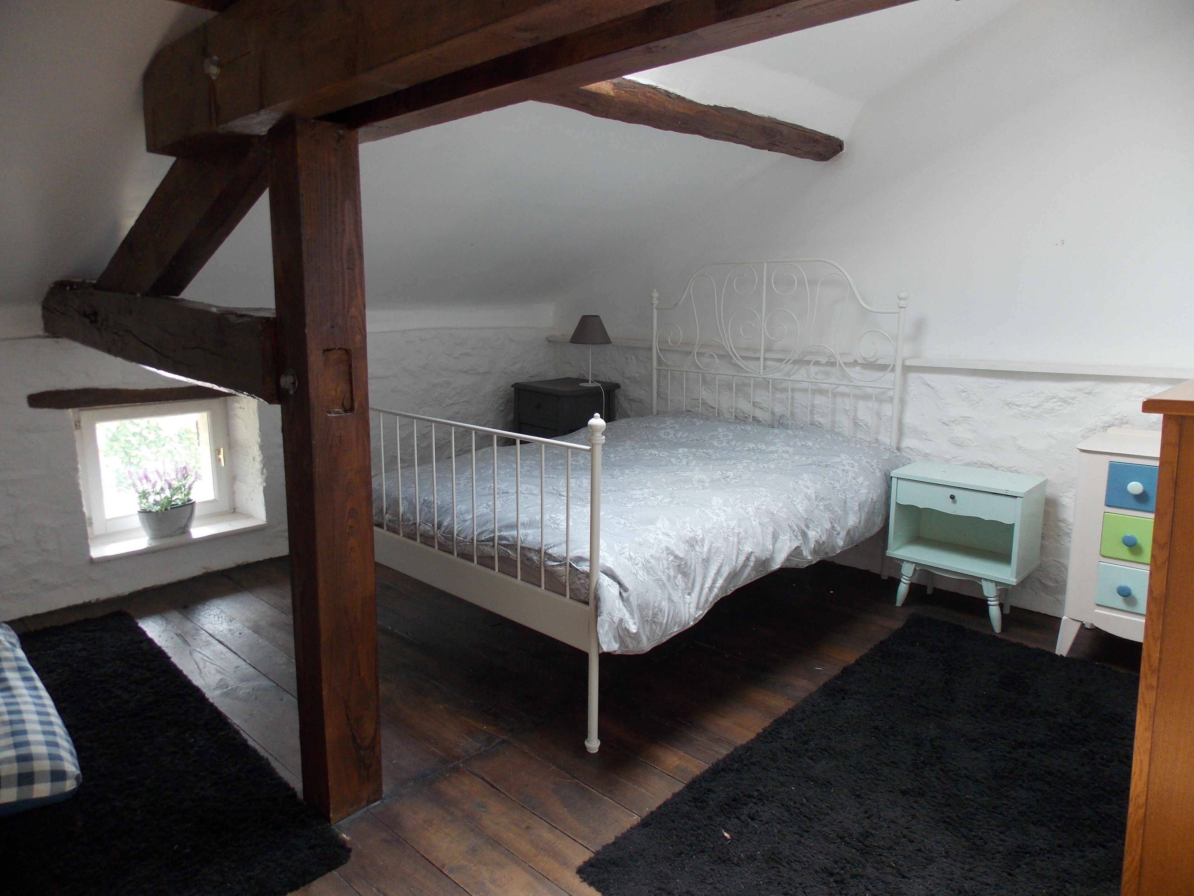 A bedroom with a single bed, a nightstand, a dresser, a window with a plant, and a wooden beam structure.