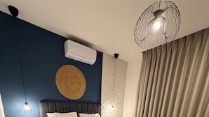 A modern bedroom with a large bed, a bedside table, an air conditioning unit, a decorative wall hanging, and a textured rug.