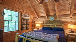 A wooden cabin bedroom with a bed, a window, a clock, and a lamp. - Secluded Log Home on 11 Forested Acres🌲🌲🌲 Jacuzzi🔥 Fire Pit🔥 Wood Stove🔥 (Dunmore)