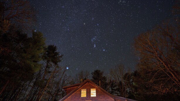 A log cabin with a starry night sky visible through the windows. - Secluded Log Home on 11 Forested Acres🌲🌲🌲 Jacuzzi🔥 Fire Pit🔥 Wood Stove🔥 (Dunmore)