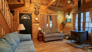 A cozy log cabin interior with a fireplace, a sofa, a small table, and a deer head mounted on the wall. - Secluded Log Home on 11 Forested Acres🌲🌲🌲 Jacuzzi🔥 Fire Pit🔥 Wood Stove🔥 (Dunmore)