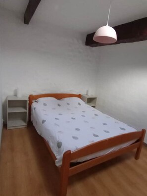 A single bed with a wooden frame and a white mattress with grey patterns. There is a white bedside table and a white shelf unit against the wall. A white pendant light hangs from the ceiling.