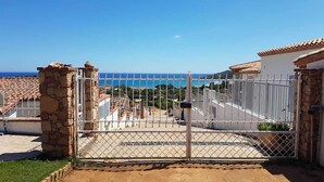 A gated entrance with a stone pillar, a metal gate, and a view of the sea and buildings in the background.