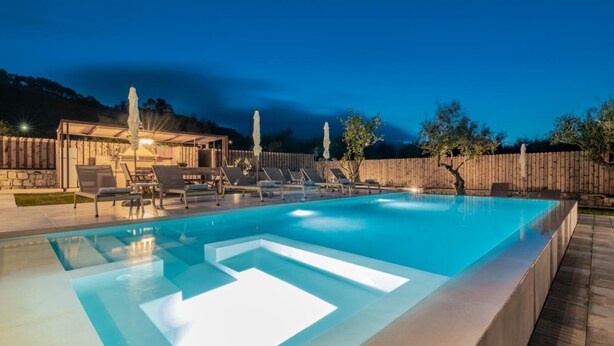 A rectangular swimming pool with a clear blue water feature, surrounded by lounge chairs and a wooden deck. - Claudio Stone House!! (Zakynthos)