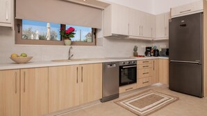 A modern kitchen with light wood cabinets, a stainless steel refrigerator, a built-in oven, and a window with a view of trees. - Claudio Stone House!! (Zakynthos)