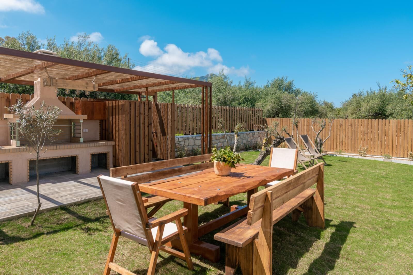 Een houten eethoek buiten met een tafel, banken, een pergola en een barbecue.