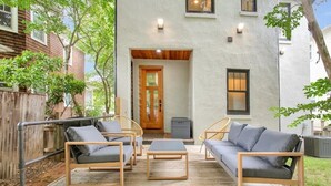 Un patio con mobiliario exterior, una terraza de madera y un edificio de fachada blanca y puerta de madera.