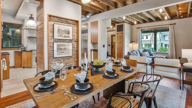 Una cocina moderna con vigas de madera, un comedor con una mesa larga dispuesta para seis personas y una sala de estar acogedora con un sofá y un ventanal grande.