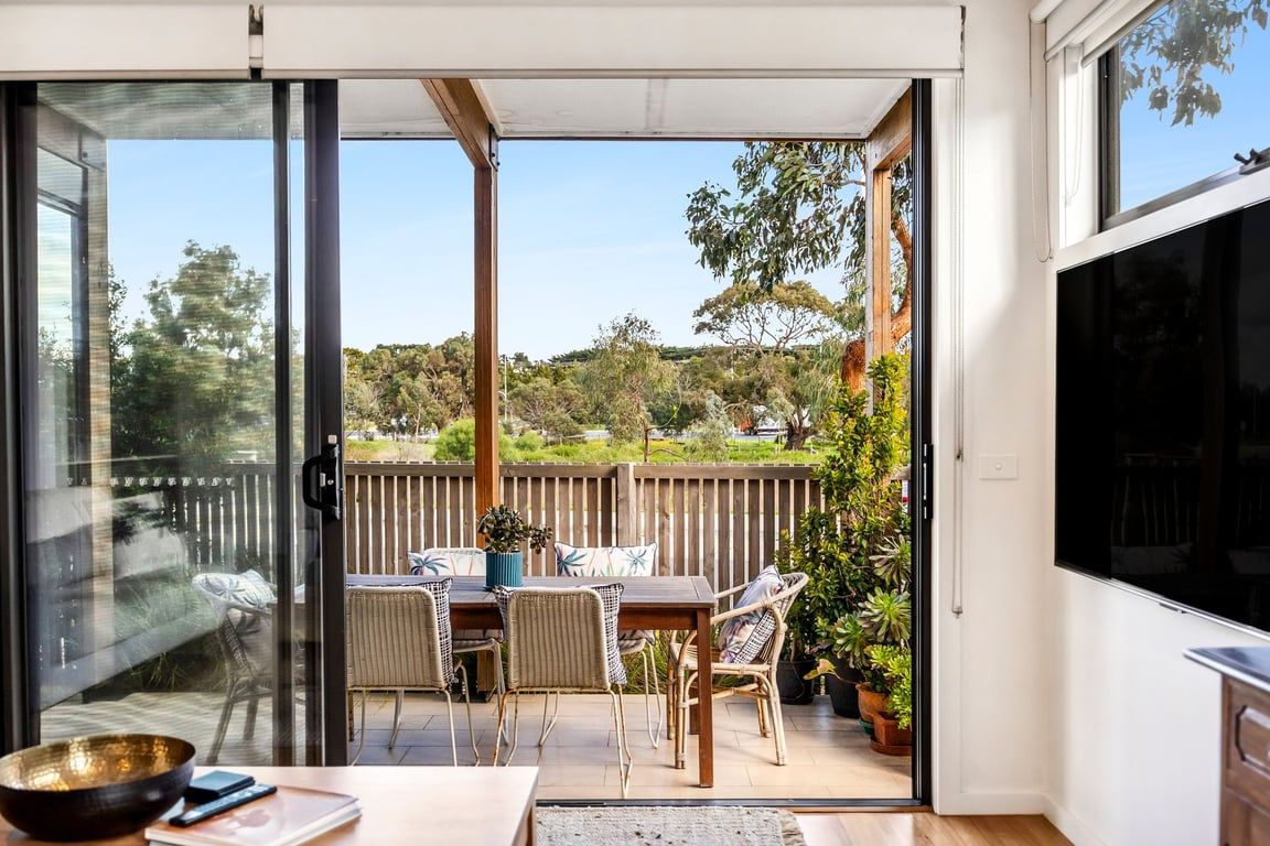 Une pièce dotée d’une porte-fenêtre coulissante donnant sur un patio extérieur avec une table et des chaises, une clôture en bois et des plantes en pot.