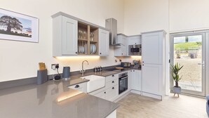 A modern kitchen with white cabinets, a large island, stainless steel appliances, and a view of a garden through the sliding glass door. - Luxury 2-Bed Cotswolds Barn with Two Ensuite Kings (Gloucester)