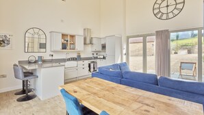A spacious kitchen with a wooden dining table and blue chairs, a large blue sofa, and a view of the outdoors through a sliding glass door. - Luxury 2-Bed Cotswolds Barn with Two Ensuite Kings (Gloucester)