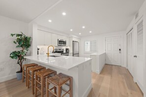 A modern kitchen with white cabinetry, a central island with stools, and a potted plant. - Loggerhead Cay #474 is a stunning 2 brm, 2 bath condo located on Sanibel! (Sanibel)