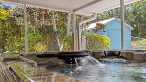 A hot tub with a waterfall feature, surrounded by lush greenery and a blue house in the background. - Tropical Tiki Hot Tub Pool Tiki Bar Game Room Pool Table 4 miles to Beach (Vero Beach)