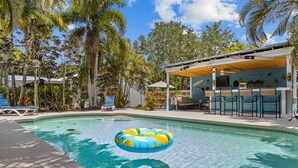 A swimming pool with a floating ring, surrounded by palm trees and a covered seating area with bar stools. - Tropical Tiki Hot Tub Pool Tiki Bar Game Room Pool Table 4 miles to Beach (Vero Beach)