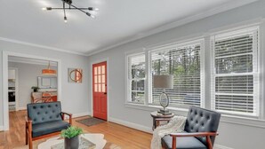 Un salón espacioso con pisos de madera, dos sillones de cuero negro, una mesa de centro blanca con una planta y una puerta roja.