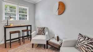 Una habitación con un sillón blanco, un sofá con cojines de estampado de cebra, un reloj de madera redondo, una mesita con un jarrón y un escritorio con taburetes.