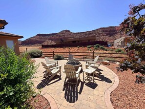 Um pátio com cadeiras Adirondack e uma lareira, cercado por um jardim paisagístico com vista para uma cordilheira.