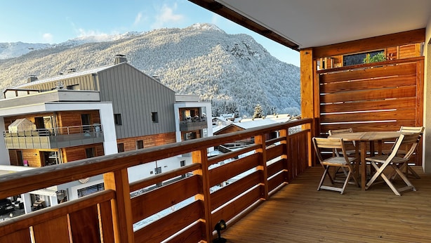 Une terrasse en bois avec une table et des chaises, d’où l’on peut admirer une montagne enneigée et des bâtiments.