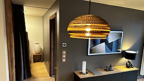 Une chambre d’hôtel moderne avec un mur gris foncé, un plancher en bois, une console noire sur laquelle se trouve un ordinateur portable, une bougie blanche et une œuvre d’art aux motifs géométriques.