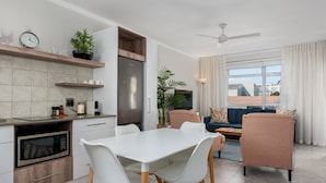 Eine moderne Küche mit integrierten Geräten, ein Essbereich mit weißen Stühlen sowie ein Wohnzimmer mit Sofa und Deckenventilator.