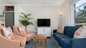 Ein Wohnzimmer mit Sofa, Sessel und Couchtisch. Ein Fernseher steht auf einem Schrank und eine Pflanze steht in der Ecke.