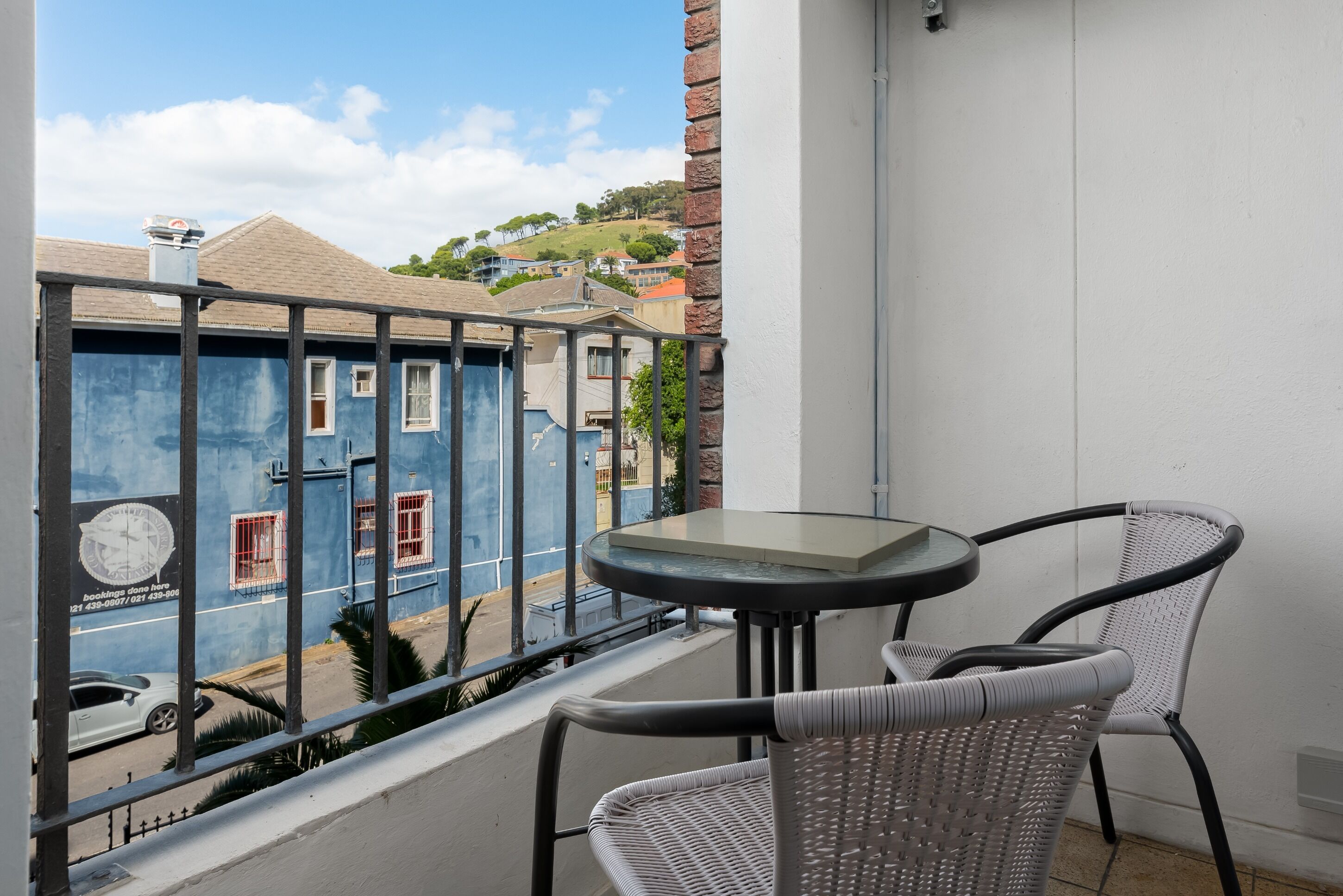 A balcony with a table and two chairs, overlooking a street with buildings and a hill in the background.