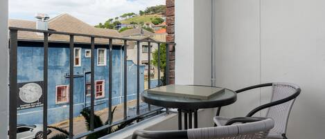 A balcony with a table and two chairs, overlooking a street with buildings and a hill in the background.